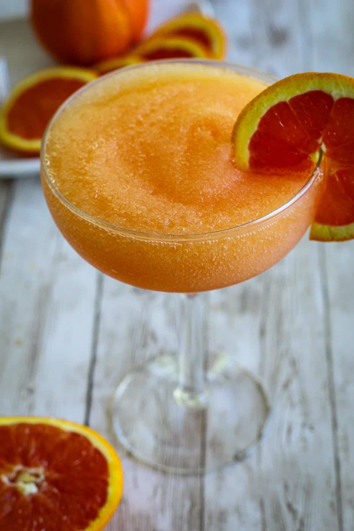 A coupe glass with a frozen drink in orange color, garnished with a slice of lemon. 