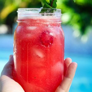 A mason jar with raspberry lemonade, being held on a hand. Setting is outdoors on a sunny day, in front of a pool.