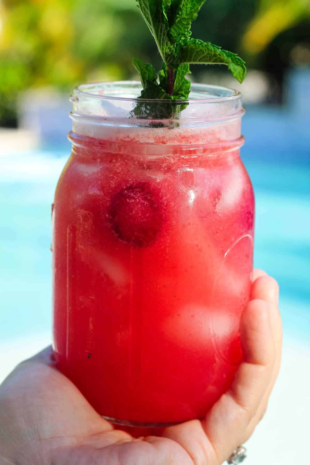 A mason jar with raspberry lemonade, garnished with fresh mint. Drink is pictured right in the center but in the background you can see a blurred pool. 