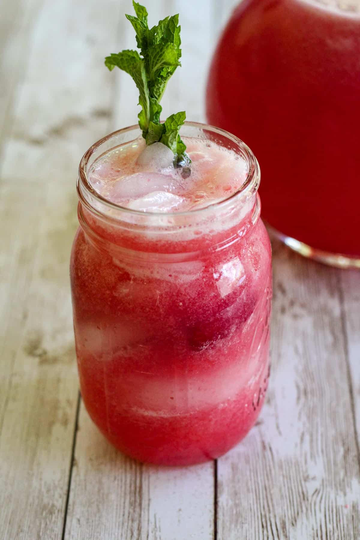 Sparkling raspberry lemonade on a mason jar, next to a pitcher with lemonade. 