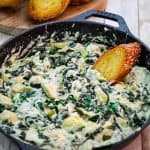 A cast iron skillet showing a dip with spinach , artichoke and goat cheese and a slice of toasted baguette dipped in it.