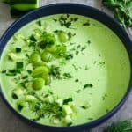 A soup plate with a green soup, garnished with chopped cucumbers, chopped mint and dill.