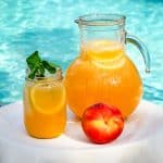 A table with white tablecloth by the pool showing a pitcher of peach lemonade, a jar of peach lemonade garnished with mint and a fresh peach too.