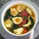 A soup bowl with tortellini soup where you can see sausage slices, spinach, carrots in a clear broth.