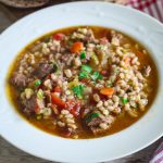 A plate with soup that shows veggies, beef and barley. Soup is garnished with parsley.