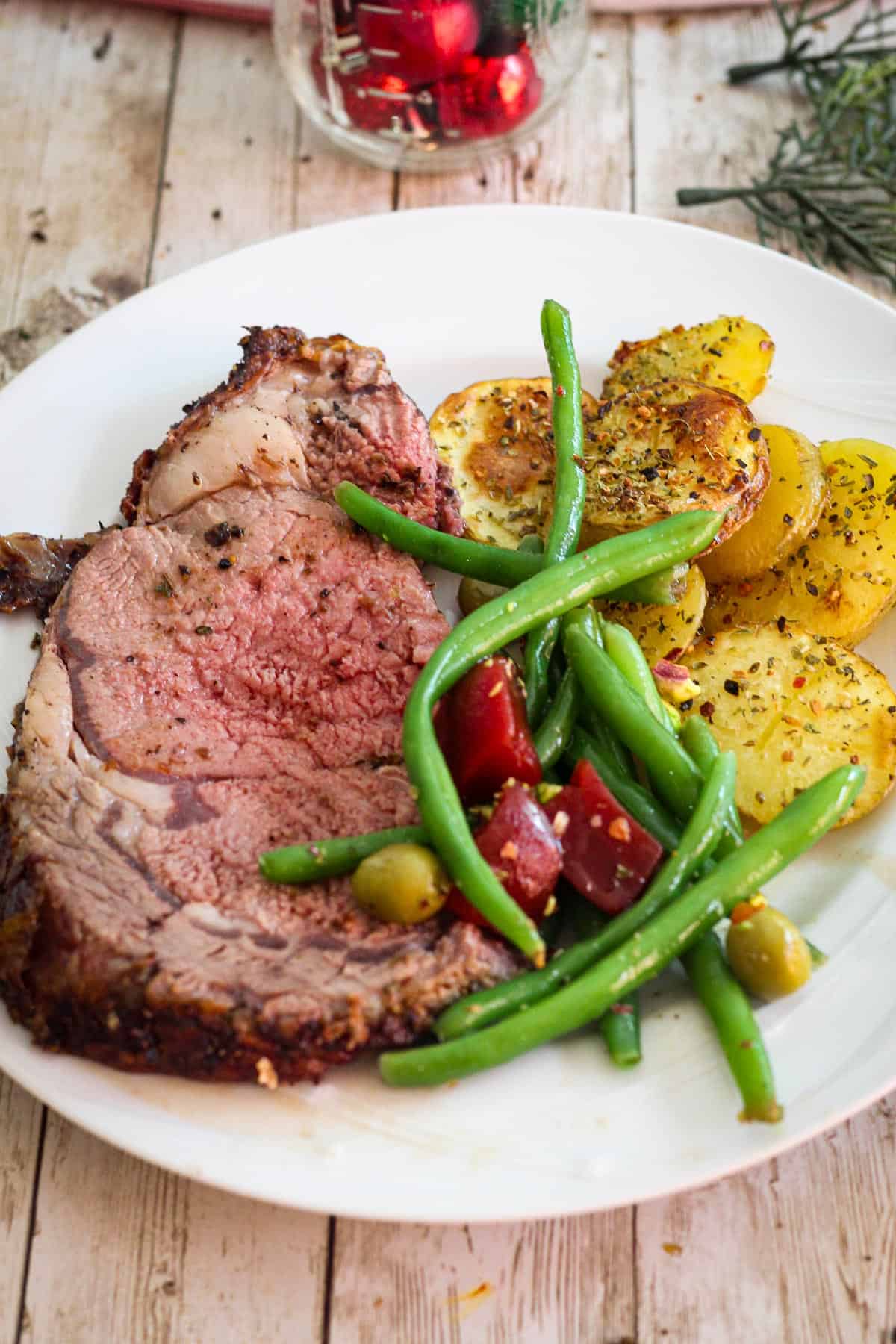 Serving of prime rib on a white plate with roasted potatoes and green beans salad. 