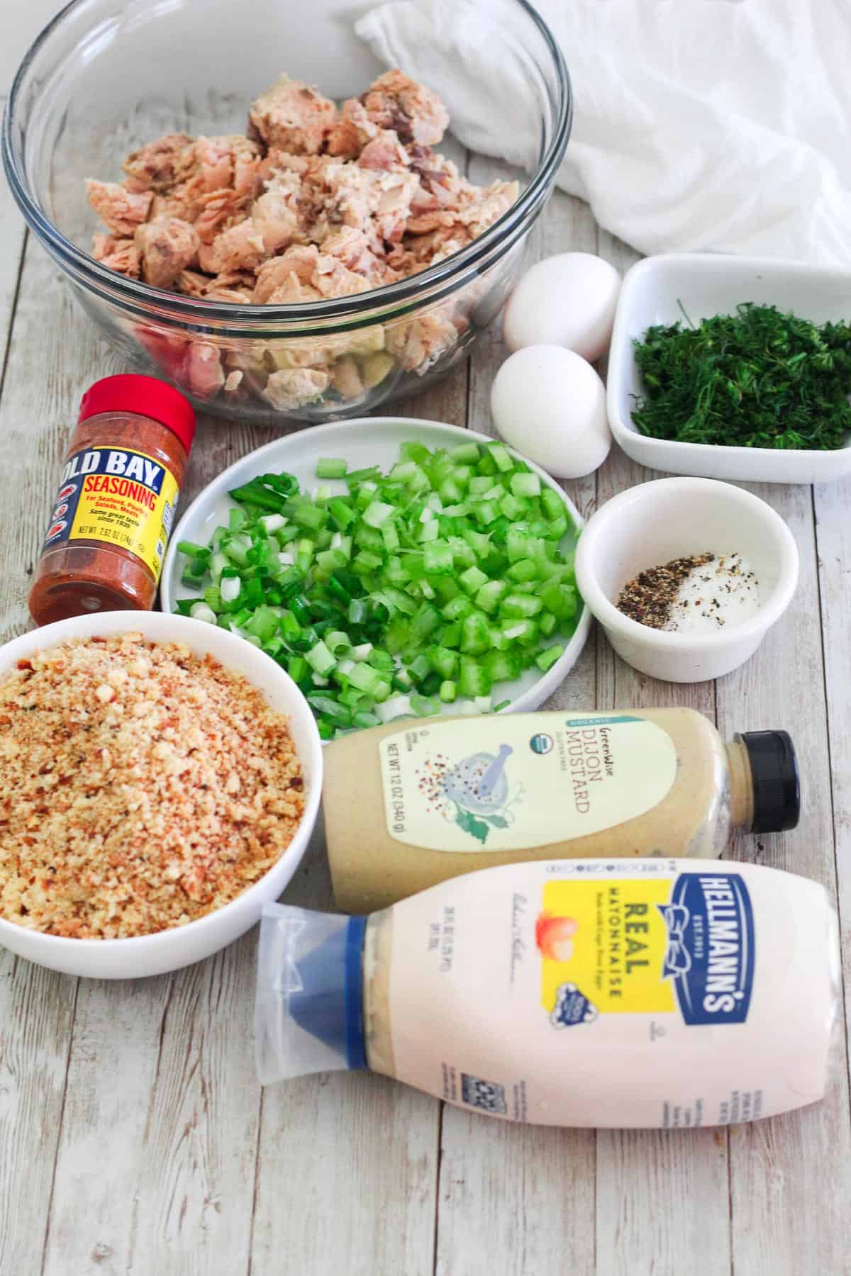 Ingredients on a counter for making salmon cakes: salmon on a glass bowl, eggs, dill, chopped celery and scallions, salt, pepper, old bay seasoning, breadcrumbs, dijon mustard and mayonnaise. 