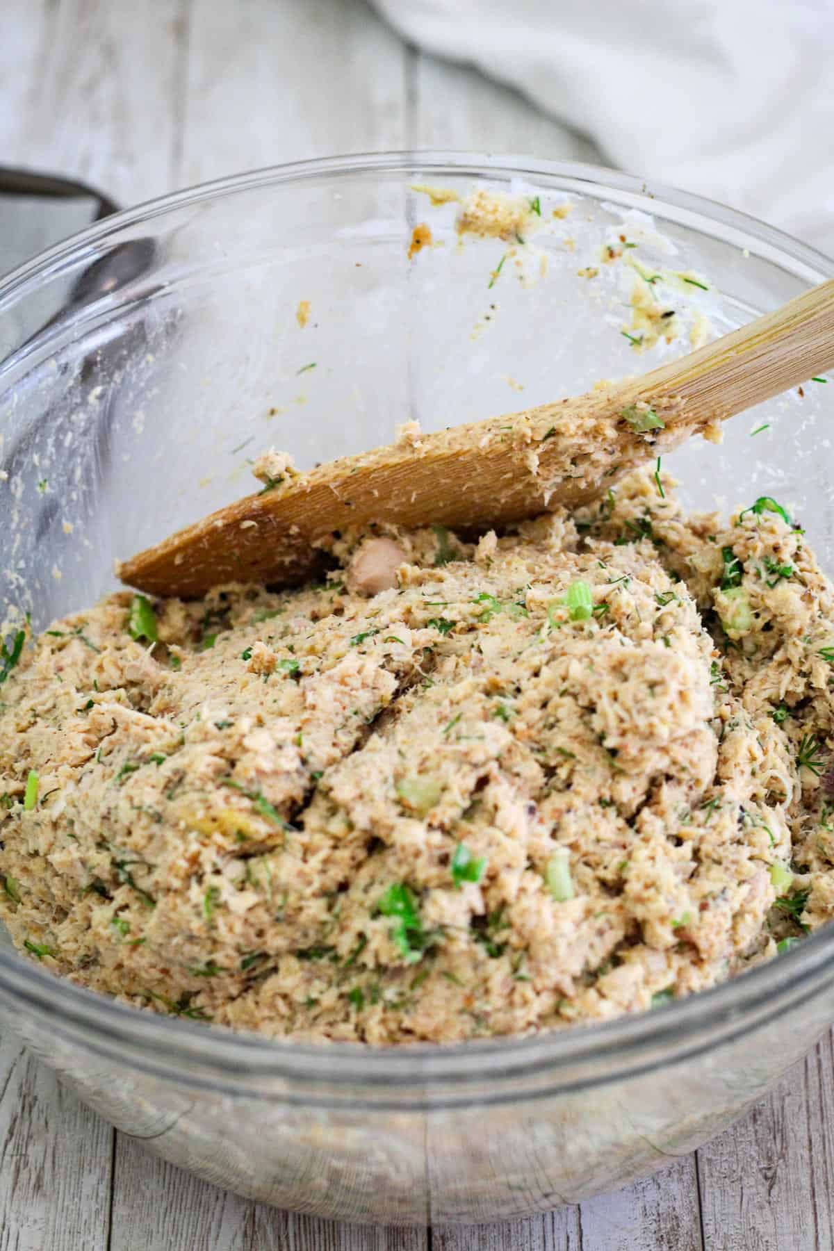 Dough for salmon cakes is all mixed and shown in the glass bowl with a wooden spoon. 