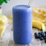 A blue smoothie in a glass, surrounded by fresh fruits in the background like bananas and blueberries.