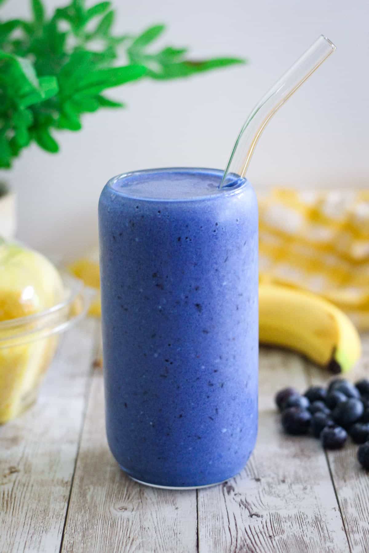 A blue smoothie with fruits and coconut water in a glass jar, surrounded by pineapple chunks, bananas and blueberries. 