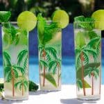 Three tall glasses by the pool with mojitos in them. Glasses are garnished with mint and limes.