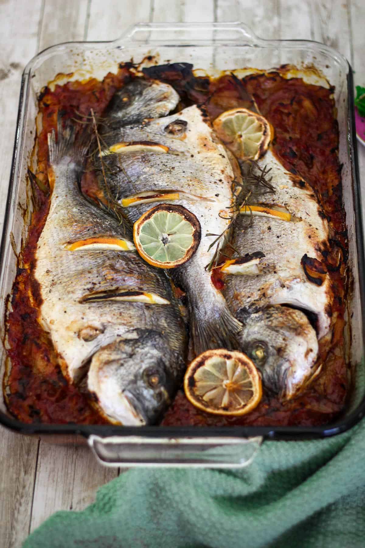 Baked sea bream fish in a baking dish. Baking dish has a layer of sauce with three fish on top, garnished with lemon slices.