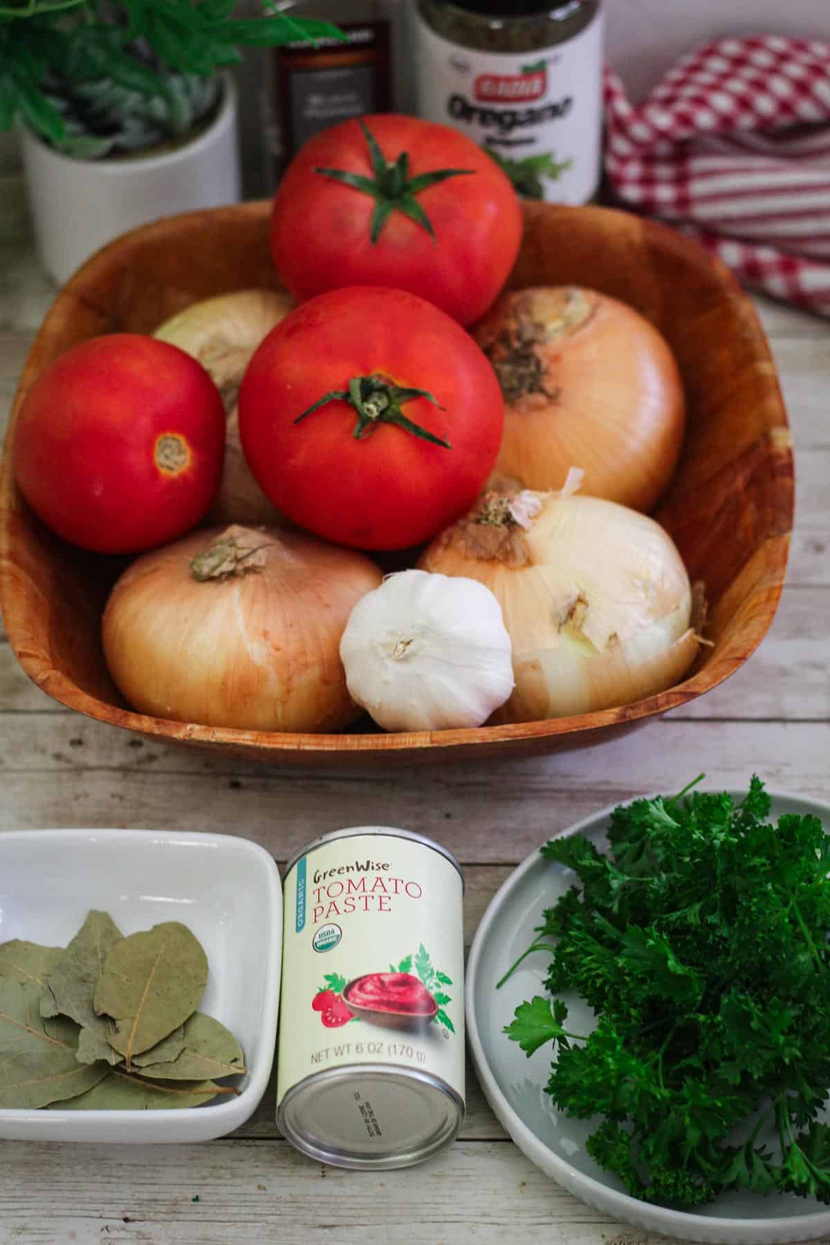 Onions, tomatoes and garlic on a basket shown next to bay leaves, tomato paste can, parsley and oregano. 