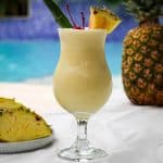 A pina colada drink by the pool, shown next to some real pineapple and garnished with cherries and pineapple.