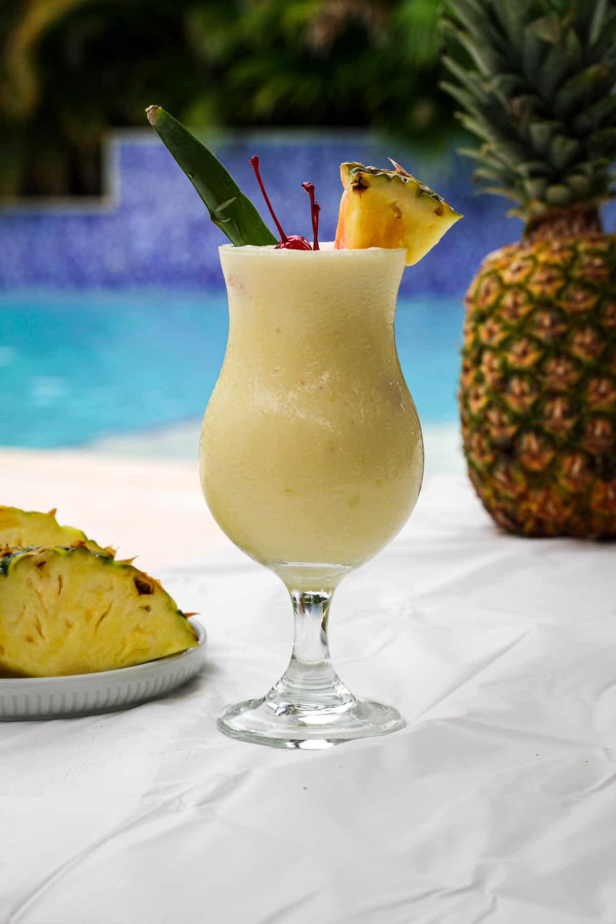 A piña colada drink on a table by the pool. There's a real pineapple in the background and more slices of pineapple close to the drink.