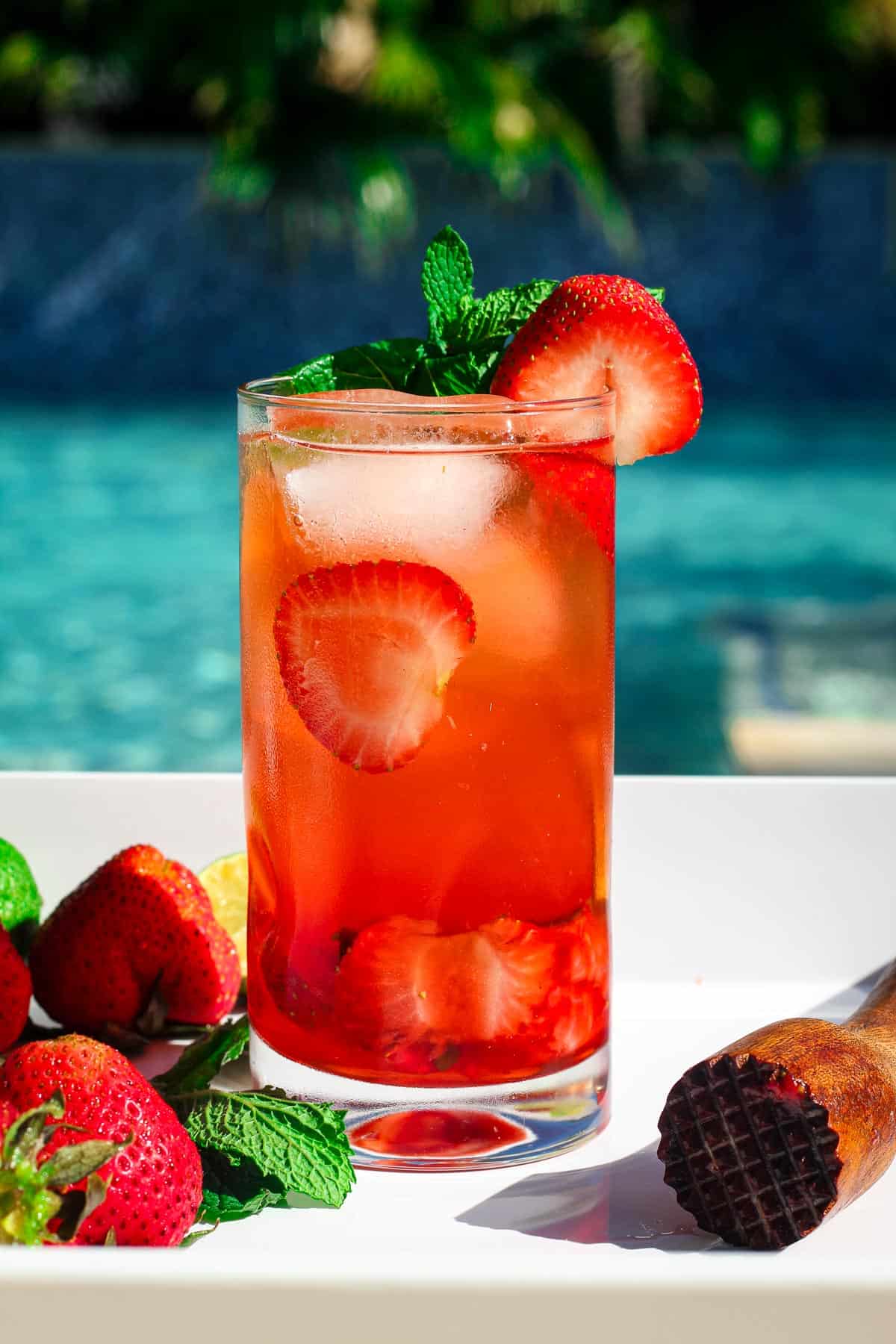 A strawberry mojito tall glass garnished with fresh strawberries and mint leaves, shown poolside. 