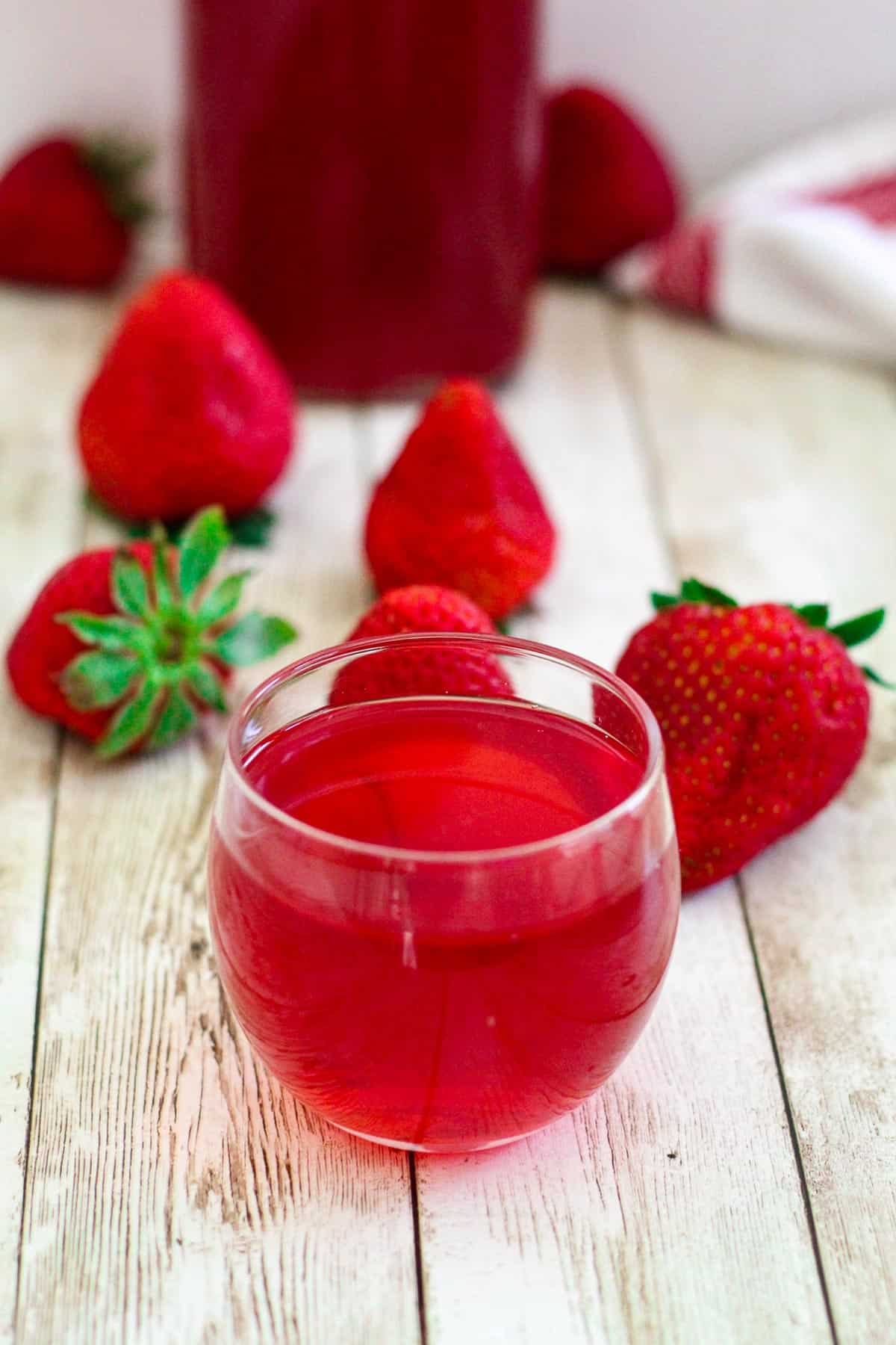 Strawberry simple syrup shown in a small glass cup next to fresh strawberries. In the background there's a whole bottle of this syrup. 