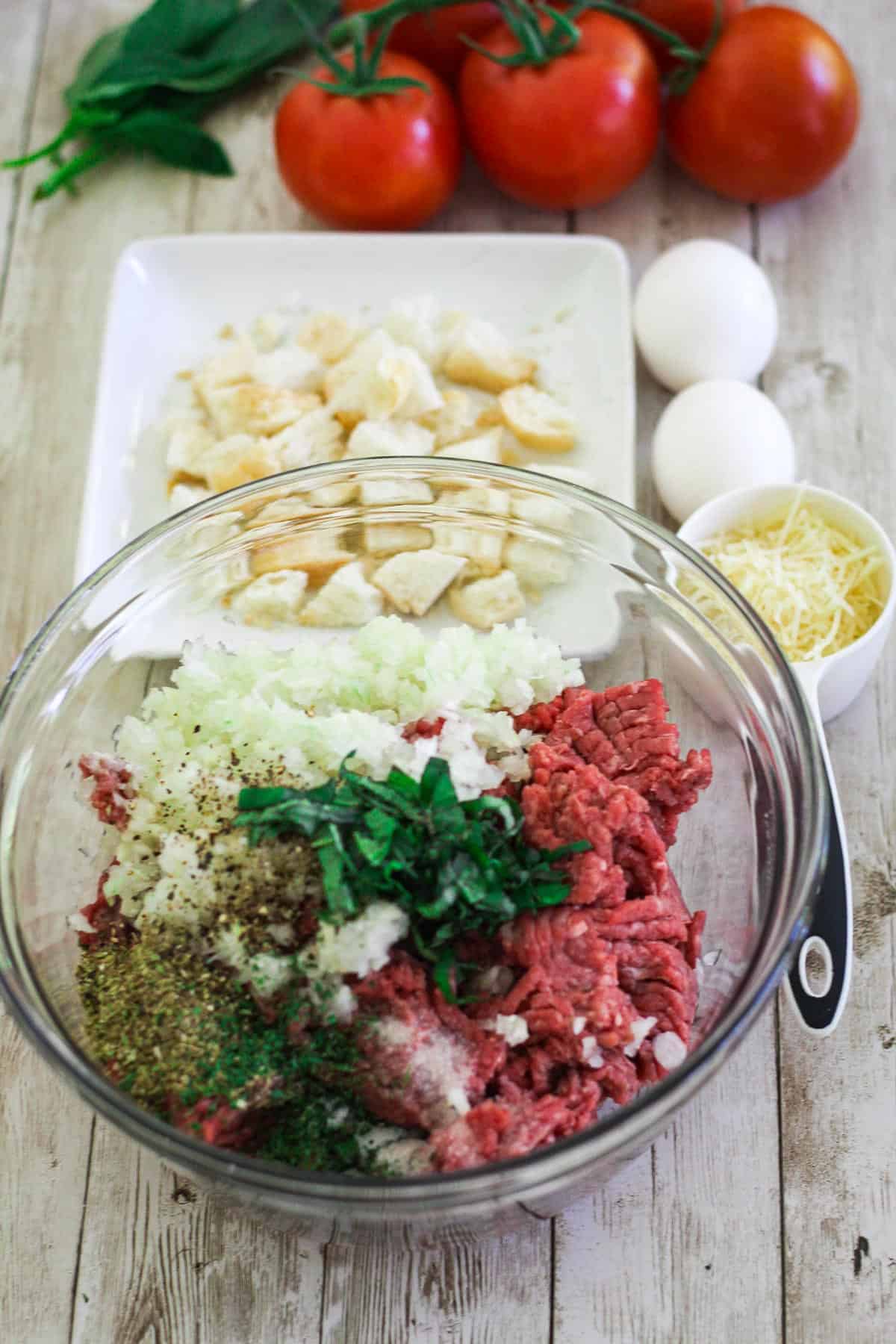A glass bowl showing most ingredients to make meatballs: ground beef, seasonings, onions, fresh basil etc. On the side of the bowl there's parmesan cheese, eggs, stale bread etc etc. 