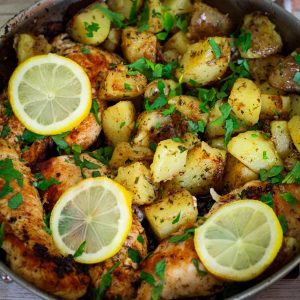 Skillet with cooked chicken and potatoes, garnished with parsley and lemon slices.