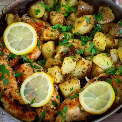 Skillet with cooked chicken and potatoes, garnished with parsley and lemon slices.