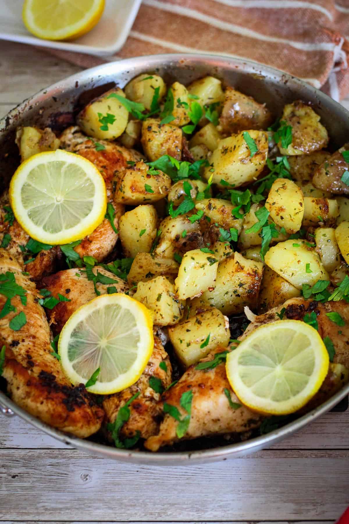 A stainless steel skillet showing cooked chicken and potatoes, garnished with lemon slices and fresh parsley. 