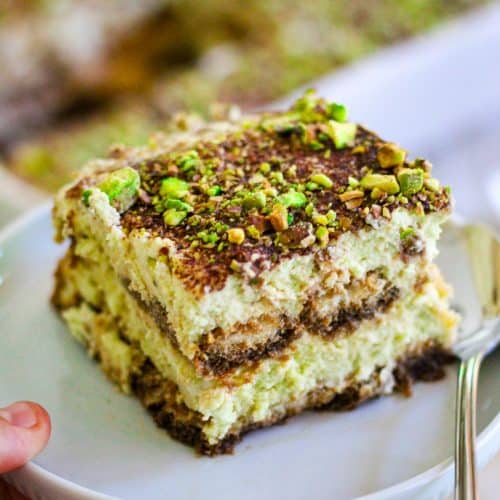 A slice of pistachio tiramisu on a plate, with the tray of tiramisu in the background.