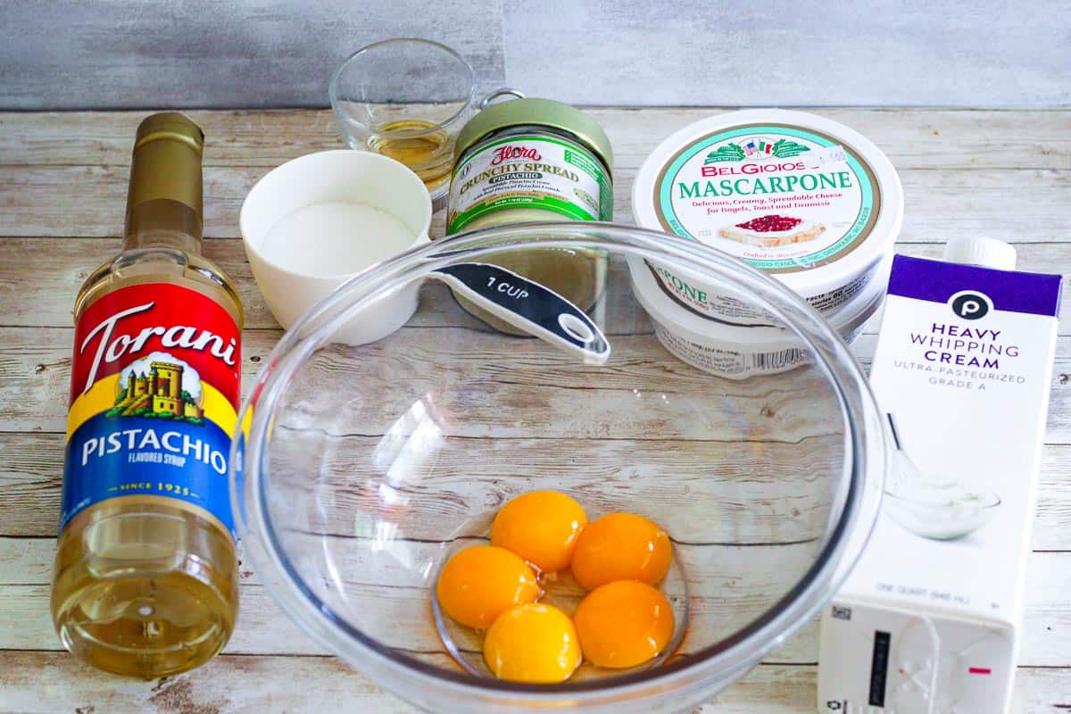 Some ingredients on a counter for making pistachio tiramisu. Visible from left to right: pistachio syrup, sugar, amaretto, pistachio cream, mascarpone, egg yolks, heavy cream. 