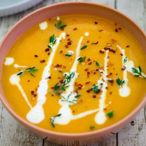 A bowl of butternut squash and red pepper soup, served garnished with cream, thyme and red pepper flakes.