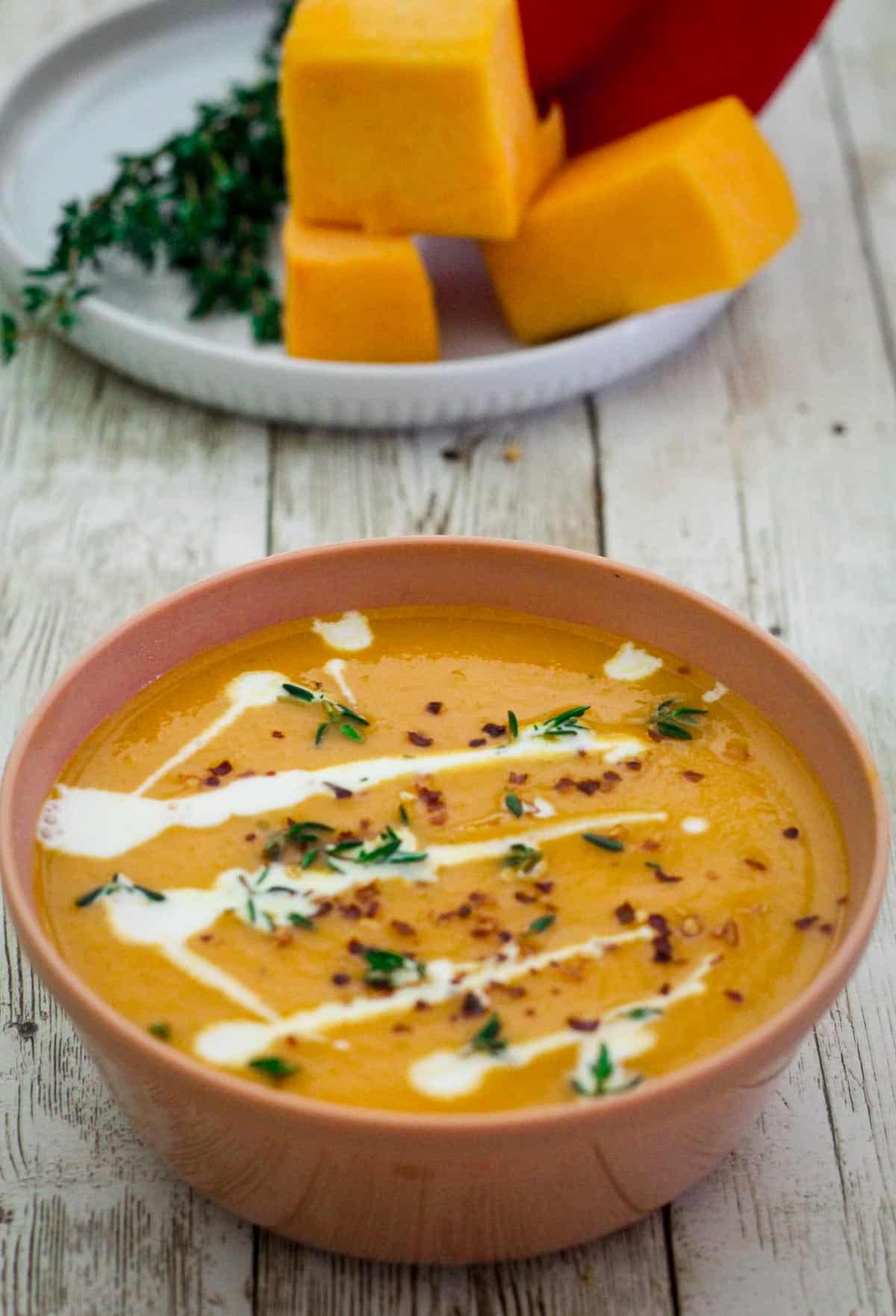 A bowl of creamy soup garnished with cream, thyme and red pepper flakes. In the background there's fresh cubes of butternut squash next to a red pepper. 