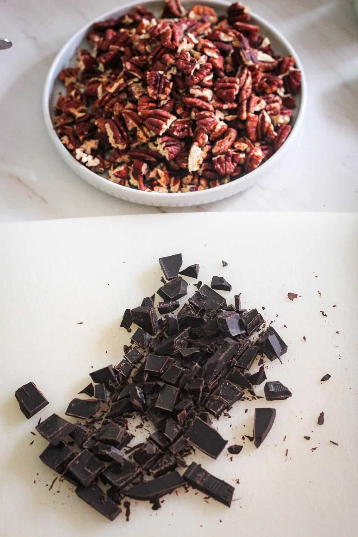 Toasted pecans on a plate in the back, while in the front there's a cutting board with chopped dark chocolate.