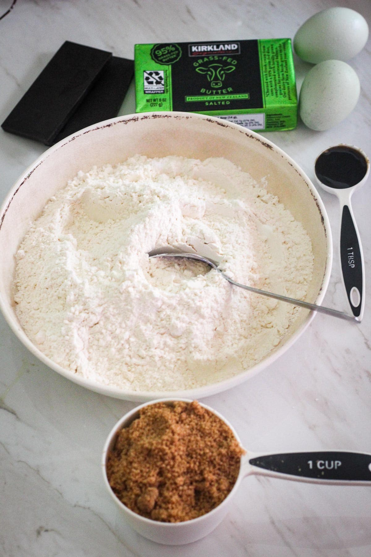 Ingredients on a counter to make cookies: chocolate, butter, eggs, vanilla paste, flour, brown sugar.