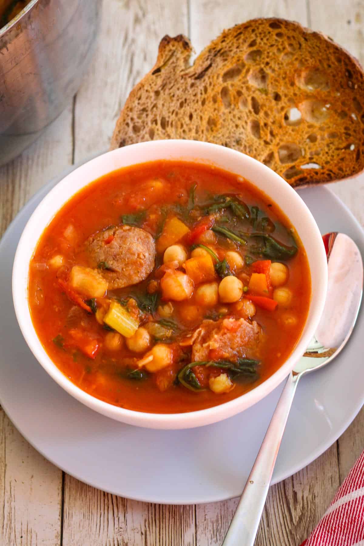 Bowl of chickpeas soup with sausage and spinach. It's a tomato based soup and it's served with a slice of toasted bread on the side.