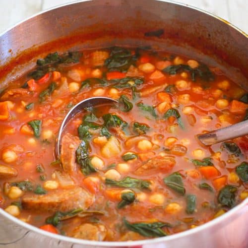 A pot of chickpea soup with Italian sausage and spinach. There's a ladle in the soup.