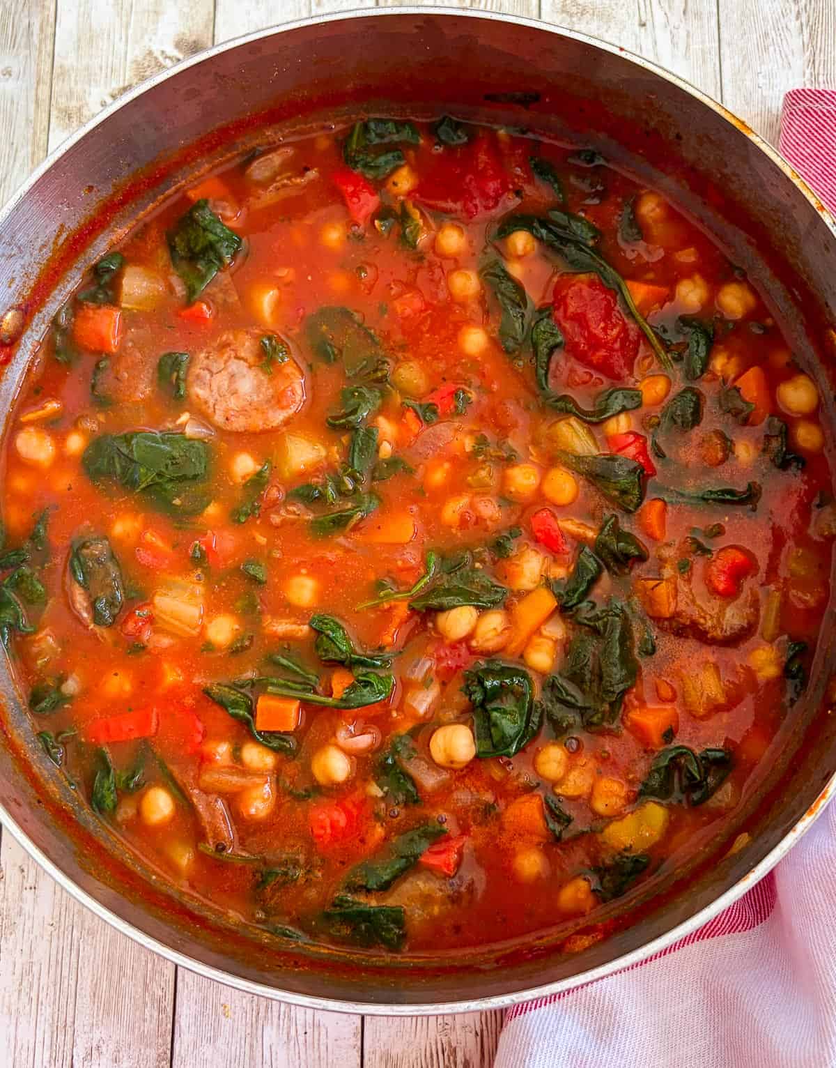 Cooked chickpeas soup with sausage and spinach in a soup pot, shown from above.