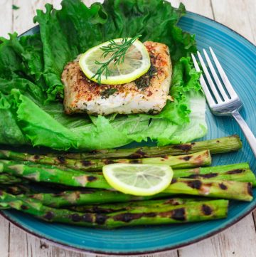 A plate with pan seared mahi mahi fillet, topped with lemon slice and dill. Served over lettuce and next to grilled asparagus.