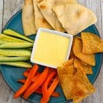 A round plate showcasing a yellow sauce (sumac sauce) in the middle, surrounded by carrot sticks, cucumber sticks, chips, pita bread etc.