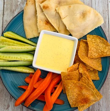 A round plate showcasing a yellow sauce (sumac sauce) in the middle, surrounded by carrot sticks, cucumber sticks, chips, pita bread etc.