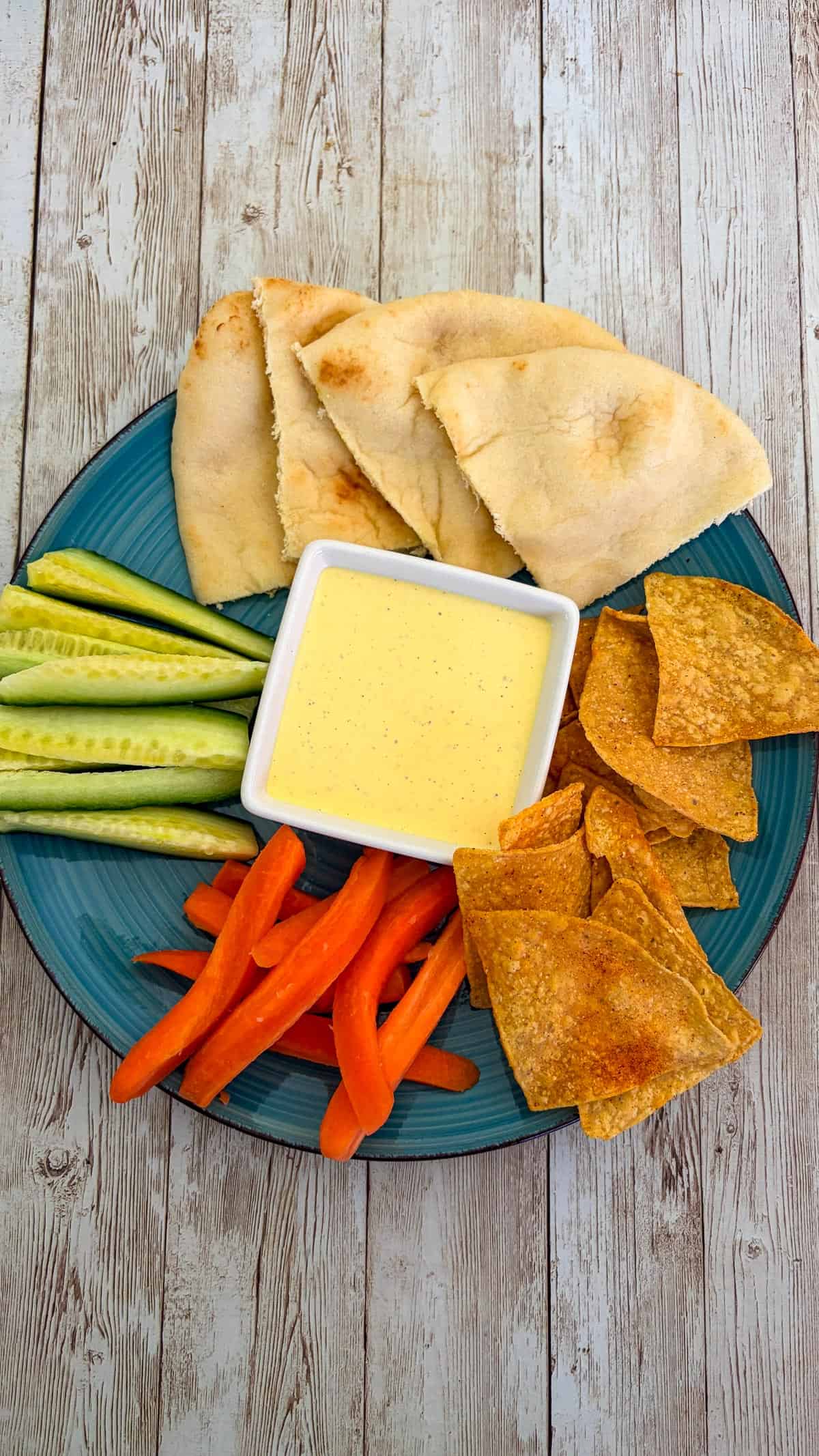 A plate showing a yellowish sauce (sumac sauce) served as a dip with veggies, pita and chips.