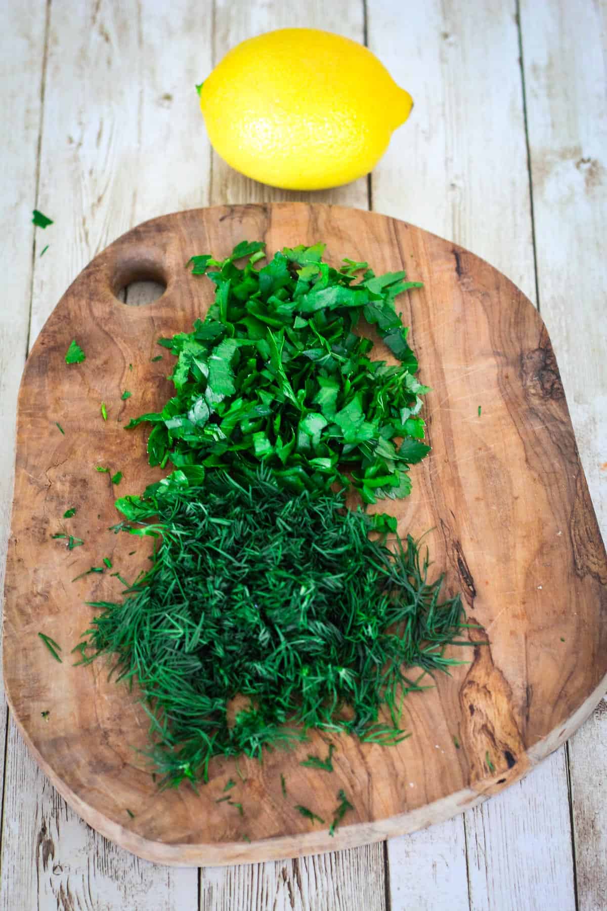 Chopped fresh parsley and chopped fresh dill on a cutting board, shown next to a lemon.