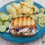 A plate with a reuben sandwich made with mahi mahi and coleslaw. Sandwich is served with chips and pickles.