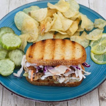 A plate with a reuben sandwich made with mahi mahi and coleslaw. Sandwich is served with chips and pickles.