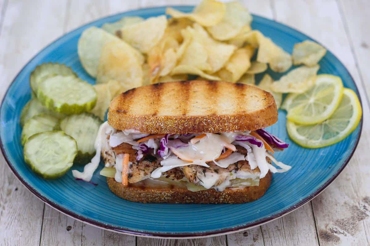 A mahi mahi reuben served on a plate with chips, pickes and lemon slices.