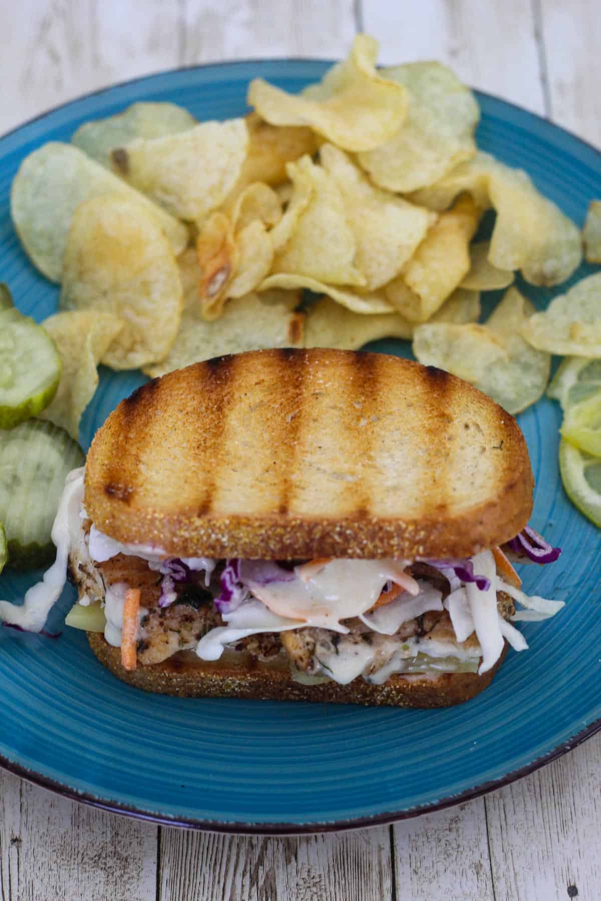 A platter with reuben mahi mahi sandwich, served with chips and pickles.