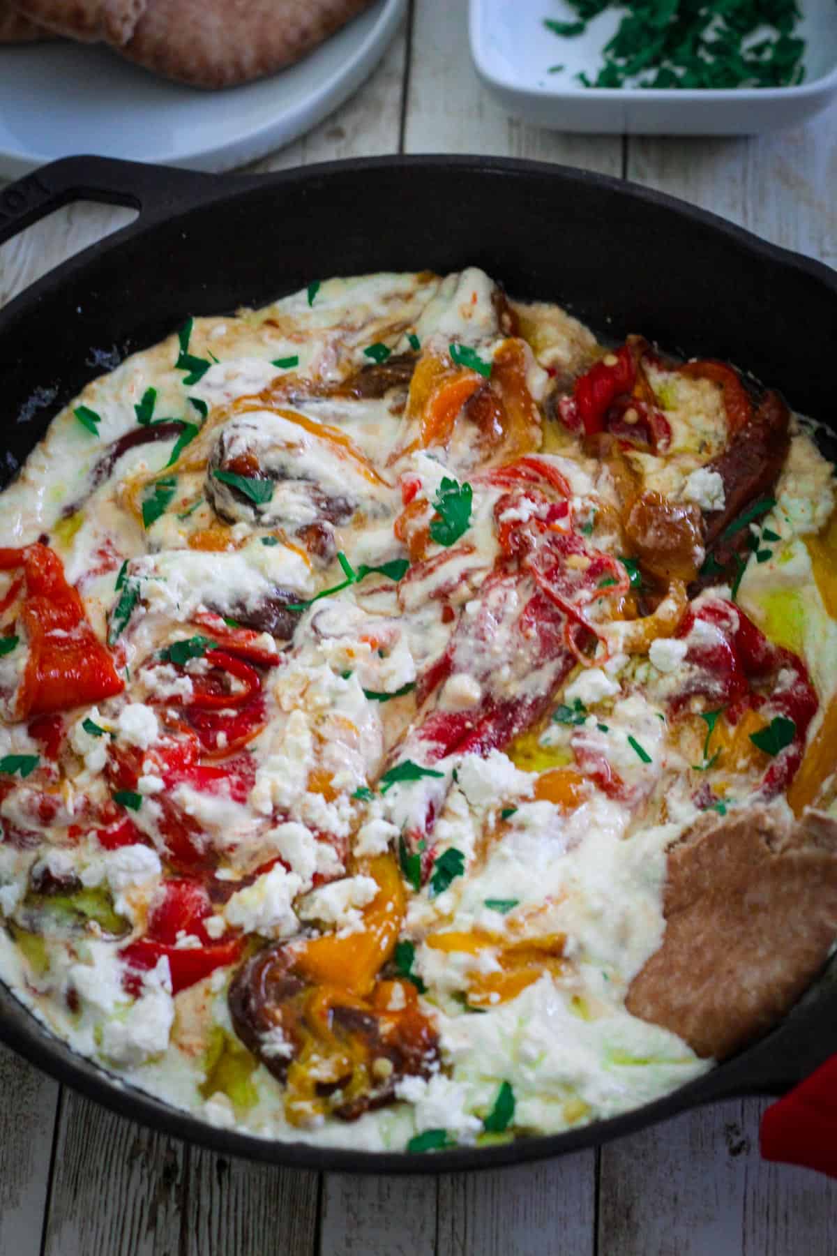 A skillet showing strips of roasted peppers mixed in a ricotta sauce. There's crumbled feta cheese over everything, with some fresh parsley and a piece of pita bread digged in.